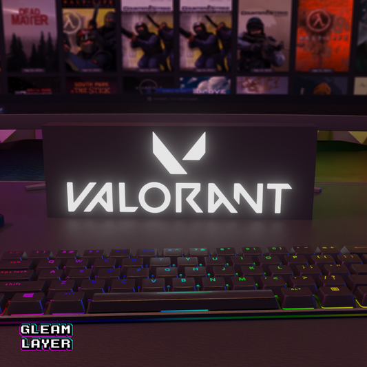 A black LED gaming light sign with the 'VALORANT' brand logo illuminated in white, placed on a desk in front of a keyboard and monitor setup.